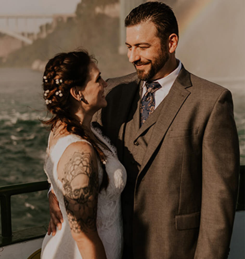 A newly married couple smiling at each other, a rainbow visible behind them 